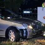 Ein beschädigter silberner Mercedes mit eingeschaltetem Scheinwerfer steht nachts am Straßenrand, daneben steht ein Mann in reflektierender Rettungskleidung mit Helm und Besen.