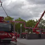 Zwei Feuerwehrkräne mit ausgefahrenen Armen auf einem Platz unter bewölktem Himmel, im Vordergrund ein Feuerwehrfahrzeug mit der Aufschrift 'FEUERWEHR'