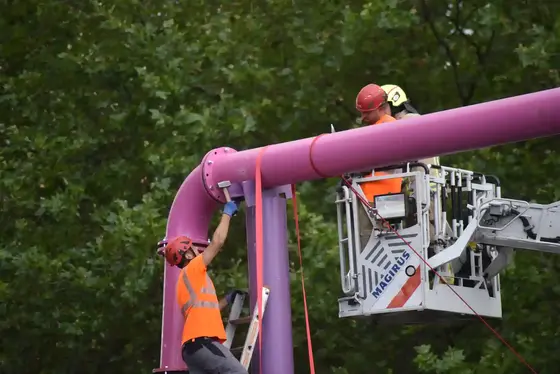 Mitarbeiter einer Fachfirma in Schutzhelmen und Warnwesten richten eine schiefe Halterung an einer großen Rohrleitung aus Metall. Im Hintergrund ein Korb einer Drehleiter.
