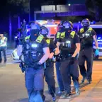 Gruppe von Polizisten in Schutzkleidung und Helmen geht nachts auf einer Straße mit Polizeifahrzeugen im Hintergrund