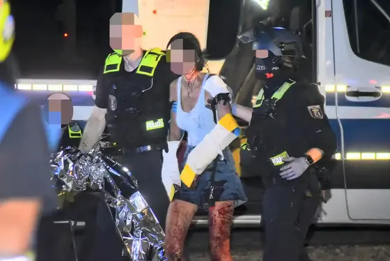 Zwei Polizisten führen eine blutverschmierte Frau mit Schiene an den Armen und blutigen Beinen an einem Fahrzeug vorbei