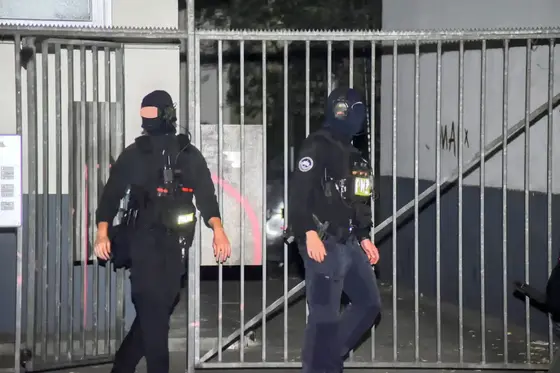 Zwei maskierte Polizisten in schwarzer Einsatzkleidung vor einem Metallzaun