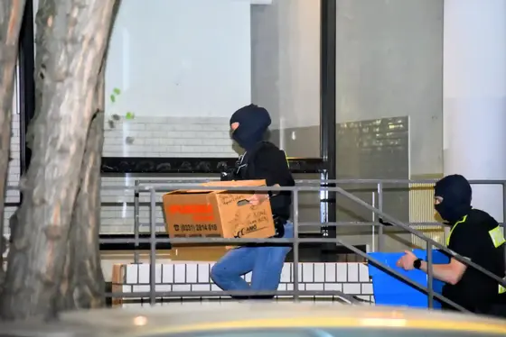 Zwei maskierte Personen tragen eine Pappschachtel und eine blaue Kiste auf einer Rampe vor einem Gebäude.