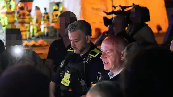 Zwei Männer, einer in Polizeiuniform mit Funkgerät und Namensschild, der andere in Anzug, umgeben von mehreren Personen und Kameraleuten in einer nächtlichen Einsatzszene.