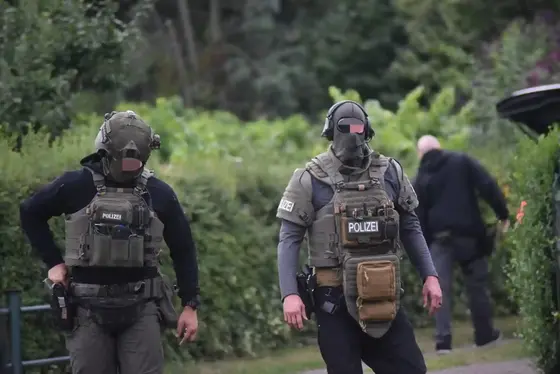 Zwei maskierte Polizisten in taktischer Ausrüstung gehen auf einem Weg, im Hintergrund eine Person neben einem geöffneten Kofferraum.