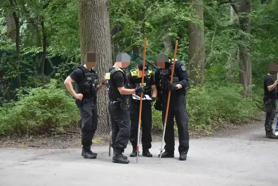 Vier Polizisten stehen auf einem Waldweg, drei von ihnen halten lange Stäbe und schauen auf eine Karte.