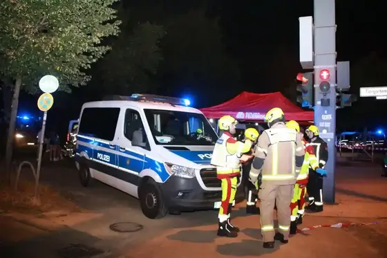 Polizeiwagen mit eingeschaltetem Blaulicht neben Feuerwehrleuten in Schutzkleidung vor einem Zelt der Verletztensammelstelle bei Nacht an einer Straßenkreuzung