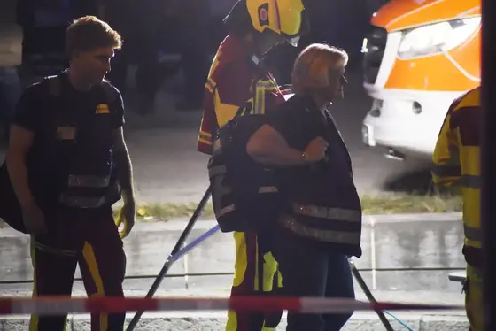 Zwei Personen in dunkler Kleidung mit reflektierenden Westen und ein Feuerwehrmann mit Helm vor einem Rettungswagen im Hintergrund bei Nacht.