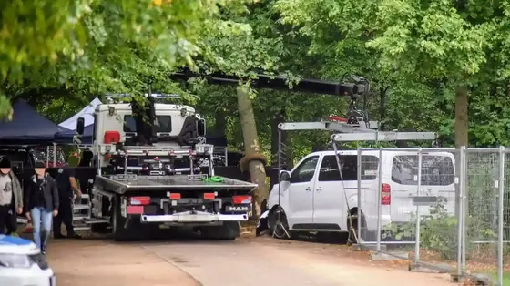 Polizeiabschleppter weißer Transporter neben einem Abschleppwagen in einem von Bäumen gesäumten Parkweg mit Bauzaun und mehreren Personen im Hintergrund