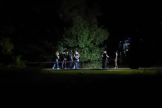 Mehrere Personen in dunkler Kleidung stehen nachts hinter einer Absperrung aus Flatterband vor einem großen Busch, rechts ist ein Fahrzeug mit Scheinwerferlicht zu sehen