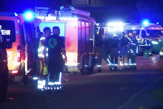 Mehrere Feuerwehrleute stehen nachts neben Einsatzfahrzeugen mit eingeschaltetem Blaulicht auf einer Straße.