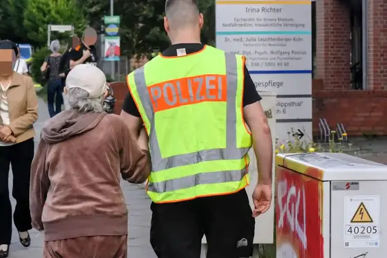 Polizist mit gelber Warnweste hält Arm einer älteren Person mit grauem Haar und brauner Kleidung auf einem Gehweg.