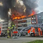 Mehrstöckiges Wohngebäude mit Flammen und schwarzem Rauch aus dem Dachgeschoss Feuerwehrleute bedienen eine Drehleiter vor dem Gebäude.