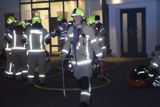 Mehrere Feuerwehrleute in reflektierender Schutzkleidung und gelben Helmen stehen vor einem Gebäude, eine Person geht mit einem Werkzeug in der Hand auf die Kamera zu.