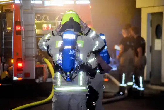 Feuerwehrmann mit Atemschutzgerät und gelbem Helm vor einem Einsatzfahrzeug, im Hintergrund weitere Einsatzkräfte und Ausrüstung bei nächtlicher Verrauchungssituation