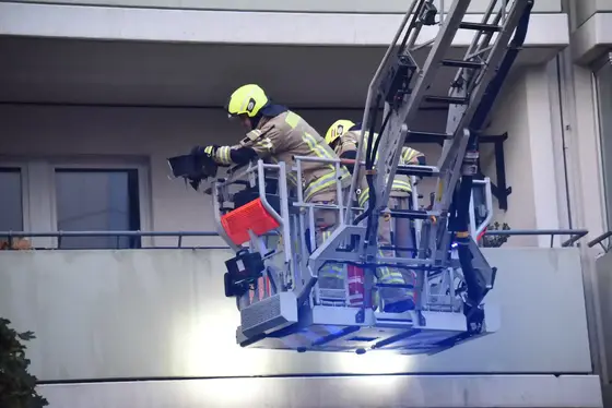 Zwei Feuerwehrleute in Schutzkleidung und Helmen stehen auf einer Drehleiter vor einem Balkon eines Gebäudes
