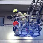Zwei Feuerwehrleute in Schutzkleidung und Helmen stehen auf einer Drehleiter vor einem Balkon eines Gebäudes