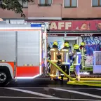 Feuerwehrleute mit gelben Helmen und reflektierender Schutzkleidung stehen vor einem Feuerwehrauto und einem Gebäude mit der Aufschrift 'MR. WAFF' und Graffiti an der Fassade