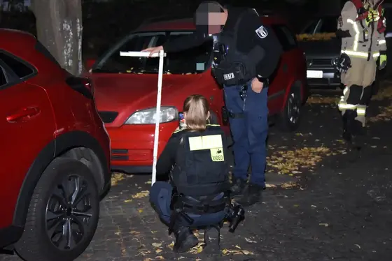 Zwei Polizisten untersuchen nachts eine Straße mit geparkten Autos, eine trägt eine Weste mit der Aufschrift 'VIDEO' und hält ein Zollstock.