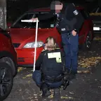 Zwei Polizisten untersuchen nachts eine Straße mit geparkten Autos, eine trägt eine Weste mit der Aufschrift 'VIDEO' und hält ein Zollstock.
