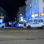 Polizeifahrzeuge und mehrere Polizisten stehen nachts auf einer Straßenecke vor einem mehrstöckigen Gebäude mit beleuchteter Kneipe