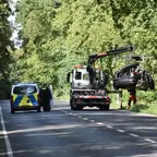 Abschleppfahrzeug hebt ein stark beschädigtes Auto an einer Straße, daneben steht ein Einsatzfahrzeug mit geöffneter Tür, umgeben von Bäumen.