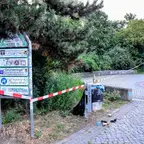 Absperrung mit rot-weißem Flatterband um eine Parkanlage mit Informationsschild und Mülleimer auf gepflastertem Weg