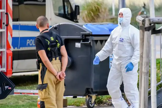 Ein Mann in Feuerwehrkleidung steht mit verschränkten Händen neben einem in weißem Schutzanzug mit blauen Handschuhen gekleideten Kriminaltechniker vor einem Müllcontainer und einem Polizeifahrzeug im Hintergrund.