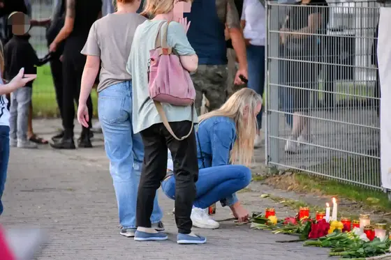 Mehrere Personen stehen an einem Zaun, eine Frau kniet und legt Kerzen und Blumen als Trauerzeichen auf den Boden.