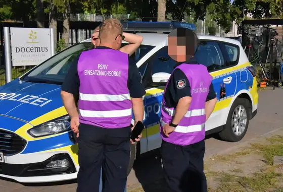 Zwei Personen mit lila Westen mit der Aufschrift 'PSNV EINSATZNACHSORGE' stehen vor einem Polizeiauto mit blau-gelber Beklebung.