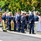 Gruppe von Feuerwehrleuten und weiteren Personen steht in einer Reihe vor einem Schulgelände mit Blumen und Kerzen.