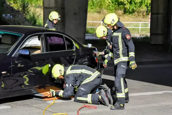 Vier Feuerwehrleute in Schutzkleidung und Helm arbeiten an einem schwarzen Auto unter einer Brücke