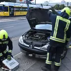 Drei Feuerwehrleute in Schutzkleidung arbeiten an einem schwarzen Peugeot mit geöffneter Motorhaube am Straßenrand.