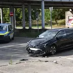 Schwarzer Kombi mit beschädigter Front steht auf Kreuzung neben Polizeifahrzeug unter Brücke