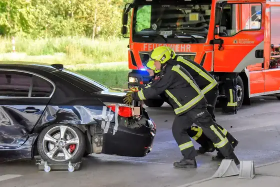 Einsatzkräfte der Feuerwehr schieben einen stark beschädigten schwarzen BMW auf einer Straße vor einem roten Feuerwehrfahrzeug