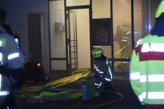 Rauch steigt im Eingangsbereich eines Gebäudes auf, Feuerwehrleute in Schutzkleidung bereiten Schläuche und Ausrüstung vor.