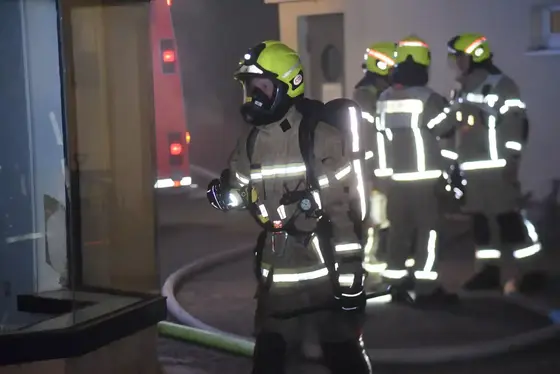 Vier Feuerwehrleute in Schutzkleidung und teils mit Atemschutzmasken stehen in einem verrauchten Hof, einer hält eine Taschenlampe, im Hintergrund ein Feuerwehrfahrzeug und Schläuche.
