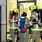 Drei Feuerwehrleute in Schutzkleidung und Helmen stehen vor einem Gebäude hinter Absperrband.