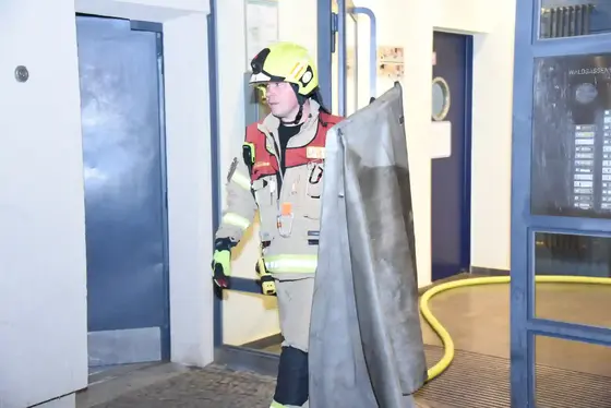 Feuerwehrmann in Schutzkleidung und Helm trägt Rauchschutzvorhang vor einem Gebäude mit offenem Eingang und gelbem Schlauch auf dem Boden.