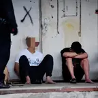 Drei Männer sitzen auf dem Boden vor einer Wand, zwei Polizisten stehen vor ihnen