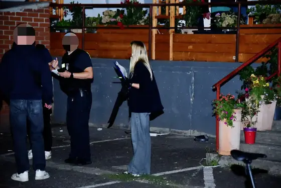 Drei Personen stehen nachts auf einem Parkplatz vor einer Backsteinmauer, eine Frau hält einen Notizblock und eine Machete und spricht mit zwei Männern, einer trägt ein schwarzes T-Shirt und eine Dienstwaffe.