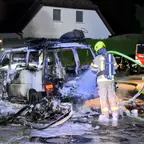Zwei Feuerwehrleute in Schutzkleidung löschen nachts einen ausgebrannten Van vor einem Wohnhaus.