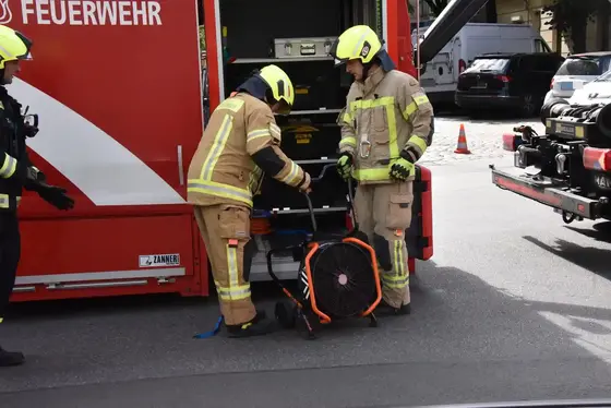 Zwei Feuerwehrleute in Schutzkleidung und Helm bedienen ein Lüftungsgerät vor einem roten Feuerwehrfahrzeug.