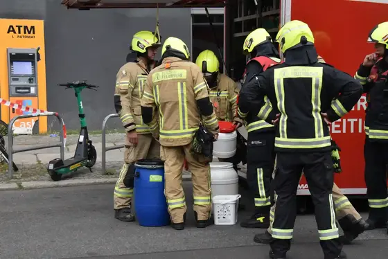 Mehrere Feuerwehrleute in Schutzkleidung und Helmen stehen um verschiedene Auffangbehälter auf einer Straße, im Hintergrund ein Geldautomat und ein E-Scooter.
