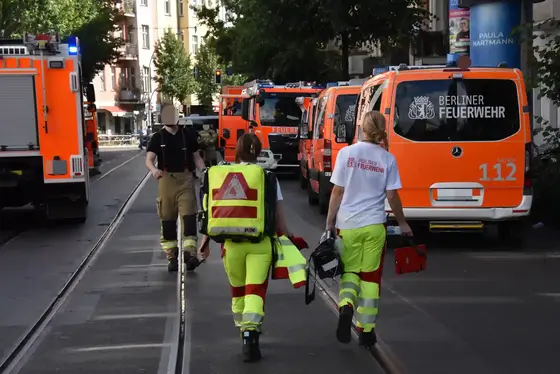 Drei Rettungskräfte in gelb-roter Einsatzkleidung und Feuerwehrmann in schwarzer Uniform gehen auf einer Straße mit mehreren orangefarbenen Einsatzfahrzeugen der Berliner Feuerwehr.