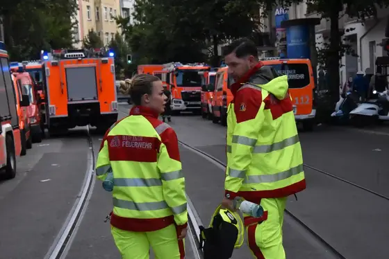 Zwei Rettungskräfte der Berliner Feuerwehr in gelb-roter Schutzkleidung stehen auf einer Straße mit mehreren Einsatzfahrzeugen im Hintergrund.
