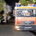 Feuerwehrfahrzeug mit der Aufschrift 'FEUERWEHR' steht auf einer Straße, im Hintergrund löschen Feuerwehrleute ein brennendes Auto.