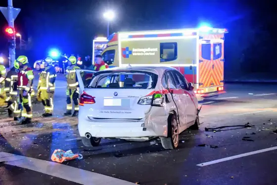 Beschädigter weißer BMW mit zertrümmerter Heckscheibe und eingedrücktem Kotflügel steht nachts auf der Straße vor einem Rettungswagen mit Blaulicht und mehreren Feuerwehrleuten in Schutzkleidung.