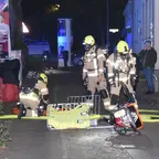 Drei Feuerwehrleute in Schutzkleidung und Helmen arbeiten nachts auf einem Gehweg mit Ausrüstung und Schläuchen, im Hintergrund stehen zwei Personen und ein Fahrrad lehnt an einer Hauswand.