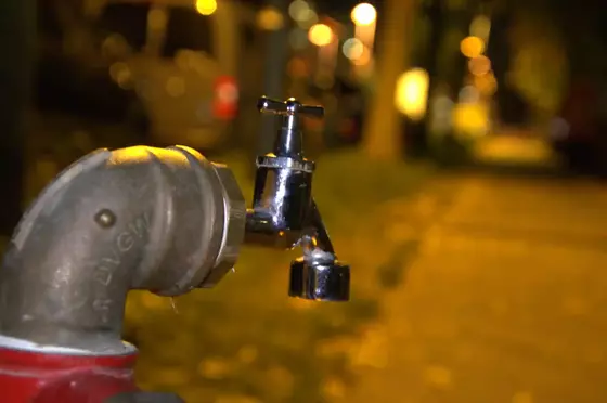 Nahaufnahme eines silbernen Wasserhahns an einem roten Rohr, unscharfer nächtlicher Straßenhintergrund mit gelben Lichtern.